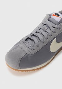 Gray athletic shoe with a combination of suede and textile upper, flat laces, a rubber outsole, and a contrasting beige swoosh logo.