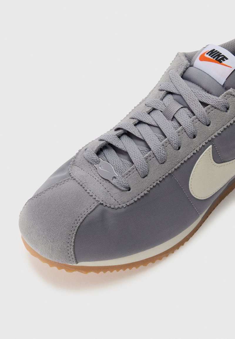 Grau sportlicher Schuh mit einer Kombination aus Veloursleder und Textil-Obermaterial, flachen Schnürsenkeln, einer Gummisohle und einem kontrastierenden beigen Swoosh-Logo.