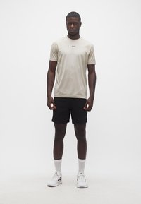 Άνδρας όρθιος, φοράει μπεζ T-shirt BOSS, μαύρα σορτς, λευκές κάλτσες και λευκά αθλητικά παπούτσια Adidas, μπροστά σε απλό λευκό φόντο.