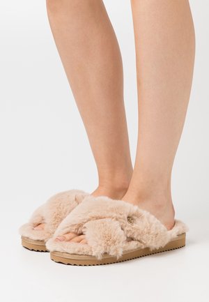 Chausson beige pelucheux avec une tige douce et moelleuse et une semelle plate texturée. Il présente un design à bout ouvert et une bride croisée pour un confort accru.