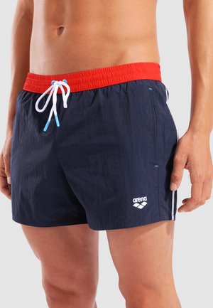 Mann trägt marineblaue Badeshorts mit rotem Bund, weißer Kordel und weißem Arena-Logo am linken Bein.