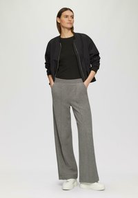 Schwarze Bomberjacke, enganliegendes schwarzes Oberteil und graue weite Hose. Glatter Stoff, lange Ärmel, Seitentaschen und weiße Sneakers.