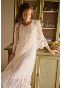 Žena v dlhých bielych čipkovaných šatách stojí vedľa rustikálneho zeleného kabinetu so sklom a košíkom s citrónmi.