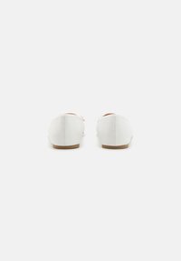 White canvas shoes with a rounded toe, flat sole, and contrasting stitching detail. The back features a smooth finish and minimal design.