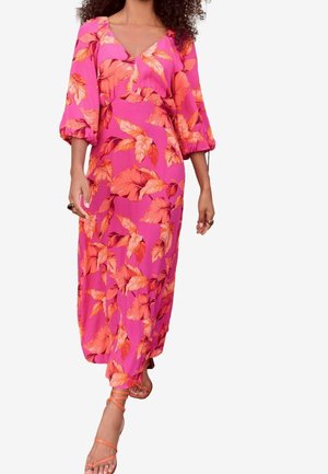 Vrouw draagt een roze maxijurk met oranje bladpatronen en opgepofte driekwartmouwen, gecombineerd met oranje sandalen met bandjes.