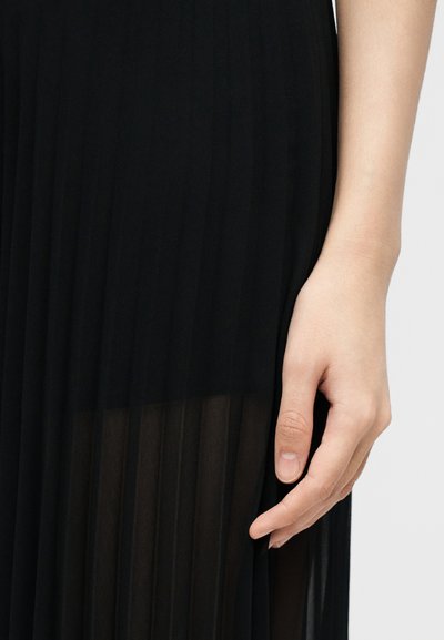 Κοντινό μιας χεριού που ακουμπάει δίπλα σε μια μαύρη φούστα με πιέτες, με διάφανο κάτω μέρος, σε ανοιχτό φόντο.