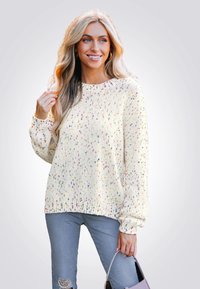 Crèmekleurige gebreide trui met veelkleurige spikkels, geribbelde halslijn en manchetten, ontspannen pasvorm. Grijze versleten jeans en een lichtpaarse tas.