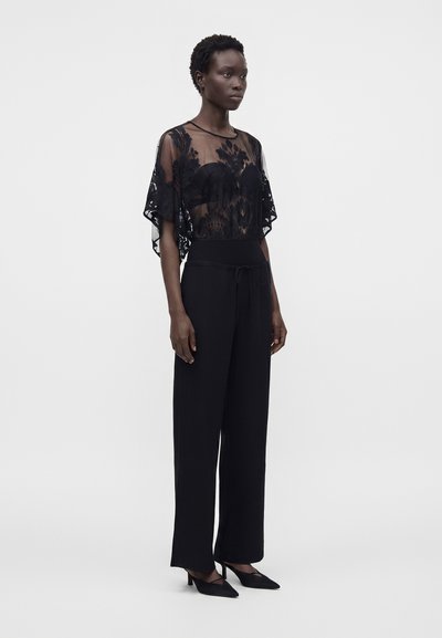 Femme debout portant un top en dentelle florale noire transparente à manches courtes, un pantalon noir taille haute à jambes larges et des talons pointus noirs.