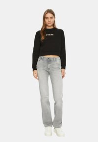 Felpa nera corta con logo "GUESS?", abbinata a jeans dritti grigio chiaro e sneakers bianche, su uno sfondo bianco neutro.