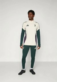 Maglietta sportiva a maniche lunghe bianca con accenti verde e viola bicolore, abbinata a leggings verdi. Presenta il marchio Adidas su entrambi i capi.