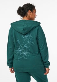 Teal huppari, jossa on kukkamallinen ääriviivadesign selkäpuolella, valmistettu pehmeästä kankaasta, pitkät hihat ja rento malli.