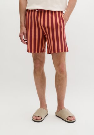 Mężczyzna w czerwono-pomarańczowych szortach w pionowe paski, beżowych klapkach z odkrytymi palcami i białej koszulce, stoi z jedną ręką w kieszeni.