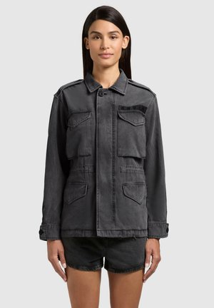 Frau mit langen dunklen Haaren, die eine dunkelgraue, geknöpfte Jacke mit mehreren Fronttaschen und schwarze Shorts vor einem schlichten Hintergrund trägt.