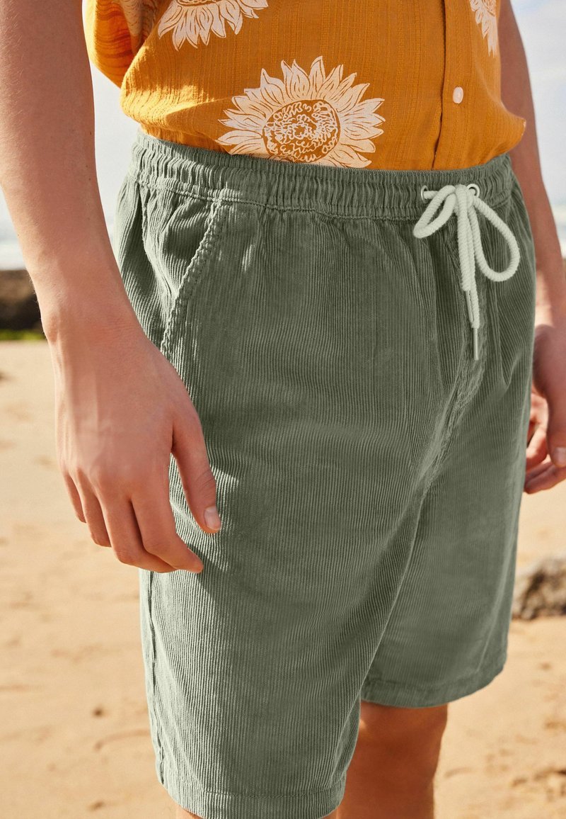 Pantalones cortos de pana verde con cintura elástica y cordón, con acanalado vertical, combinados con una camisa de botones amarilla con flores.