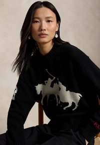 Femme aux longs cheveux noirs portant un pull noir avec un motif tricoté blanc représentant un cheval et un cavalier, assise sur une chaise en bois devant un fond uni.