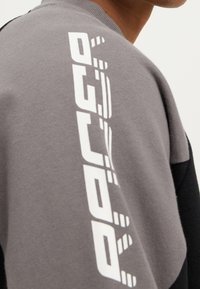 Zbliżenie na osobę w szarym i czarnym ubraniu z napisem "OUTDOOR" wydrukowanym pionowo na ramieniu w kolorze białym.