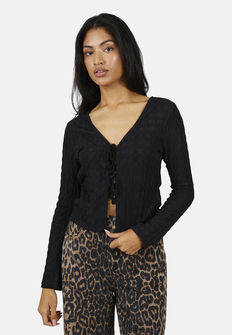 Donna che indossa una maglia nera a maniche lunghe con texture e laccetti sul davanti, abbinata a pantaloni a vita alta con stampa leopardata, in piedi su sfondo bianco.