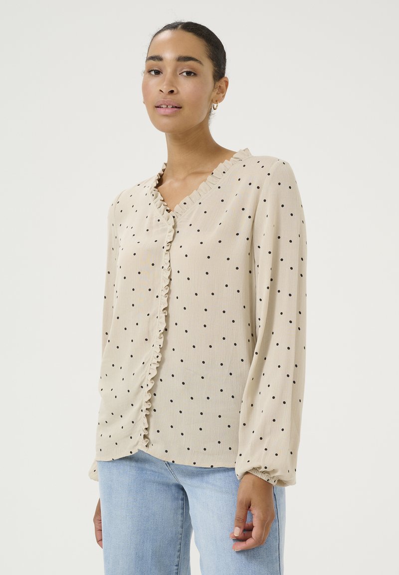 Beige blouse with black polka dots, featuring a V-neckline and ruffled trim. Long sleeves with gathered cuffs. Material appears lightweight and textured.