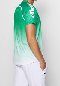 Muž v zeleno-bílé ombre sportovní košili s textem "ellesse" na rameni a bílých šortkách s černým logem, ruka v kapse, v bočním póze.