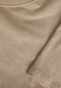 Beige katoenen T-shirt met ronde hals en korte mouwen, voorzien van een zachte textuur en subtiele stikdetails langs de zoom.