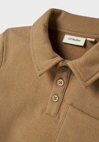 Camisa polo marrón con tres botones, un cuello y un bolsillo en el pecho, que muestra la textura de la tela y la etiqueta de la marca dentro del cuello.