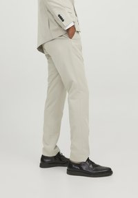 Hellbeige Anzughose mit slim Fit, glatter Textur und einer Falte an der Vorderseite. Kombiniert mit schwarzen Lederschuhen und Taschen.