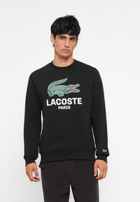 Juodas medvilninis džemperis su žaliu krokodilo logotipu ir baltu tekstu "LACOSTE PARIS". Riešo ir apykaklės kraštai su raukšlėmis.