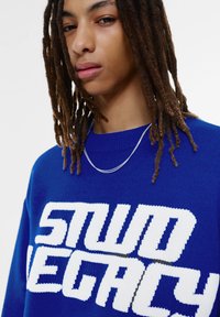 Blauwe gebreide trui met grote witte tekst "STUD LEGACY." Eenvoudig ontwerp, ronde halslijn en een gestructureerde afwerking. Simpele zilveren ketting.