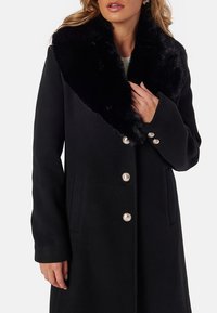 Svart ullkappa med krage av konstgjord päls, silverknappar och sidofickor. Designen har en skräddarsydd passform och långa ärmar.