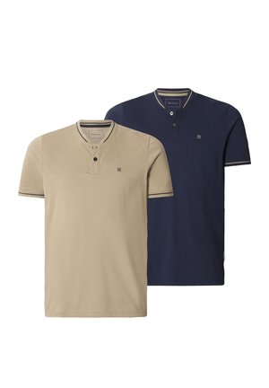 Mann trägt beige kurzärmeliges Henley-Shirt mit zwei Knöpfen und marineblauen Hosen, steht vor einem einfachen hellen Hintergrund.