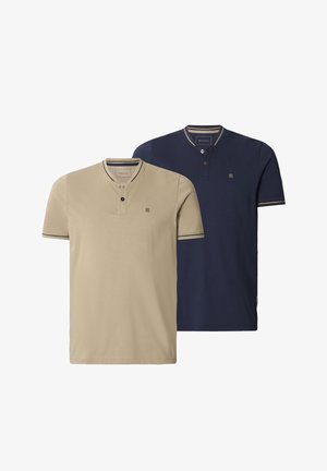 Mann trägt beige kurzärmeliges Henley-Shirt mit zwei Knöpfen und marineblauen Hosen, steht vor einem einfachen hellen Hintergrund.