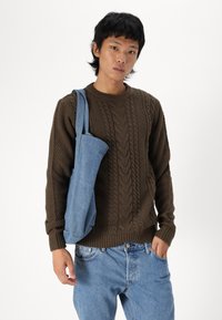 Suéter marrón de punto trenzado con dobladillo acanalado, combinado con jeans de mezclilla azul claro. Una suave bolsa tote azul cuelga del hombro.