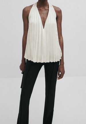 Personne portant un haut halter plissé blanc avec un décolleté plongeant en V et un pantalon noir droit sur un fond uni.