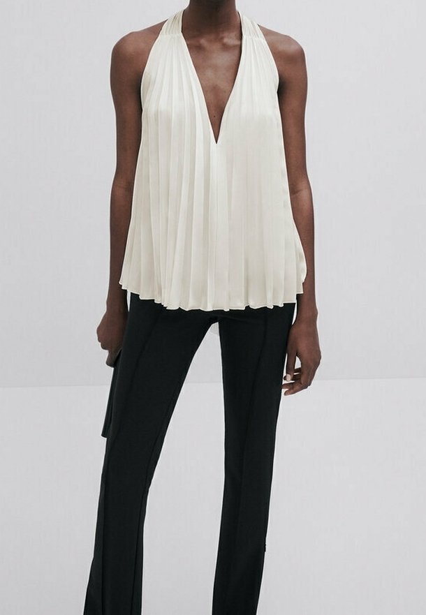 Personne portant un haut halter plissé blanc avec un décolleté plongeant en V et un pantalon noir droit sur un fond uni.