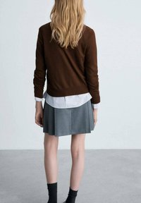 Brauner Pullover über einem weißen Hemd mit Kragen, kombiniert mit einem grauen Faltenrock. Schwarze Knöchelsocken. Die Haare sind lang und wellig. Minimalistischer Hintergrund.