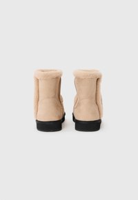 Beige suède enkellaarsjes met schapenwolvoering, afgeronde toppen en platte zwarte rubberen zolen. Eenvoudig, schoon ontwerp zonder zichtbare hardware.