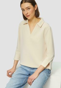 Jasno-beżowa bluzka z dekoltem w kształcie litery V, wykończona kołnierzykiem, z rękawami 3/4 i bocznymi rozcięciami. Noszona z jasnoniebieskimi dżinsami. Gładka faktura.