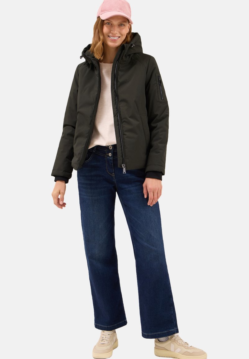 Veste rembourrée noire avec capuche, chemise légère, jeans bleu foncé à jambe large, et baskets beiges, accompagnées d'une casquette rose.