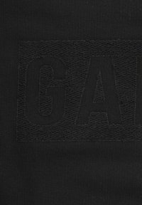 Prenda negra con tejido texturizado que presenta un logotipo "GAP" en relieve, negro sobre negro. El fondo de punto liso contrasta con la letra texturizada.