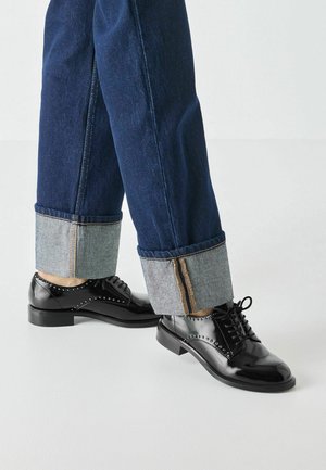 Chaussures en cuir verni noir avec des clous décoratifs, associées à un jean en denim bleu foncé avec des poignets contrastants bleu clair et des coutures visibles.