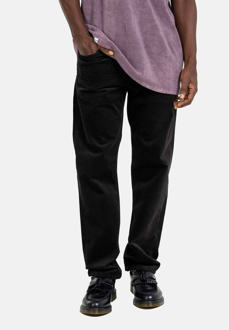 Schwarze Cordhosen mit lockerer Passform und seitlichen Taschen, kombiniert mit einem lockeren lavendelfarbenen Hemd und schwarzen Schnürschuhen mit Fransen-Details.