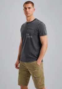 Camiseta de algodón gris con un bolsillo en el pecho y texto desgastado, combinada con un pantalón corto cargo color caqui que presenta múltiples bolsillos y un detalle de etiqueta.