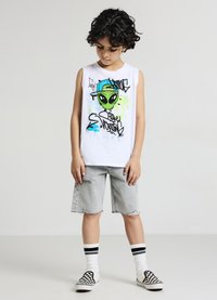 Weißes ärmelloses T-Shirt mit grünem Alien-Motiv und Graffiti-Design, kombiniert mit verwaschenen Jeansshorts. Schwarz-weiße karierte Slipper.