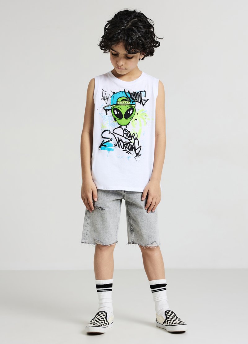 Weißes ärmelloses T-Shirt mit grünem Alien-Motiv und Graffiti-Design, kombiniert mit verwaschenen Jeansshorts. Schwarz-weiße karierte Slipper.