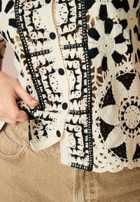 Hæklet cardigan med et blomstermønster i beige og sort, med store knapper og struktureret blondedetaljering. Accompanjeret af brune bukser.