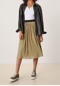 Plisserad beige kjol med svart midjeband, matchad med en vit henley-topp och svart läderjacka, avslutad med gröna sneakers.