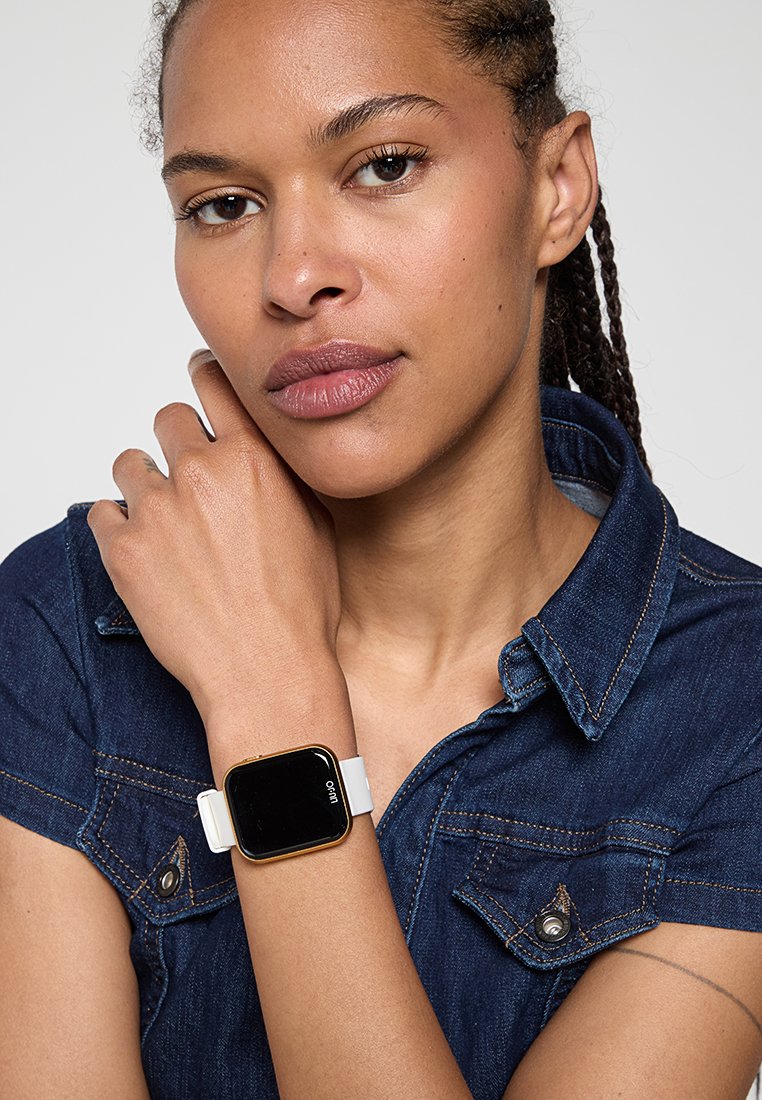 Smartwatch con uno schermo rettangolare nero e cornice dorata, dotato di un cinturino in silicone bianco. Indossato su un polso, con una camicia di jeans visibile.