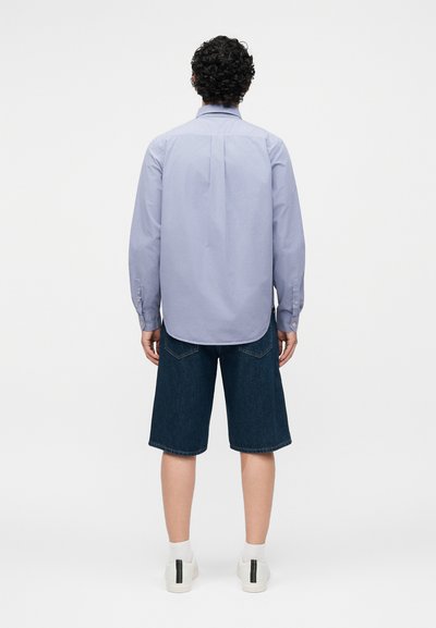 Άνδρας με κατσαρά μαλλιά, που φορά ανοιχτό μπλε πουκάμισο με μακριά μανίκια, σκούρο τζιν σορτς, λευκές κάλτσες και λευκά αθλητικά, στέκεται γυρισμένος πλάτη.