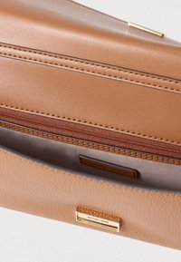 Bolso de mano de cuero marrón con una textura suave, que cuenta con un bolsillo con cremallera y un detalle de herrajes en tono dorado. El interior está forrado con una tela clara.