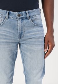 Un jean en denim bleu clair présente un design classique à cinq poches, une fermeture éclair et un bouton métallique, avec des détails de décoloration et de couture subtils.